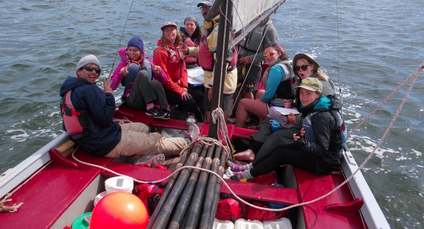 A group of people sit close together and smile at the camera while in a sailboat on choppy water. 